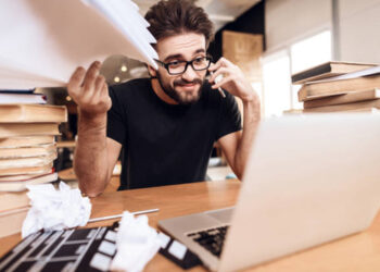 Freelancer man in t-shirt frustrated with notes at laptop sitting at desk surrounded by books.