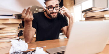 Freelancer man in t-shirt frustrated with notes at laptop sitting at desk surrounded by books.