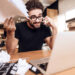 Freelancer man in t-shirt frustrated with notes at laptop sitting at desk surrounded by books.