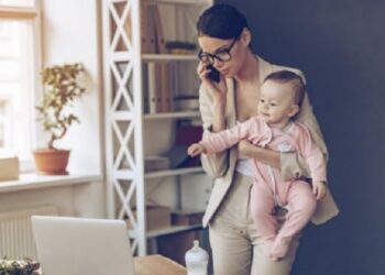 Young beautiful businesswoman talking on mobile phone and looking at laptop while standing with her baby girl at her working place