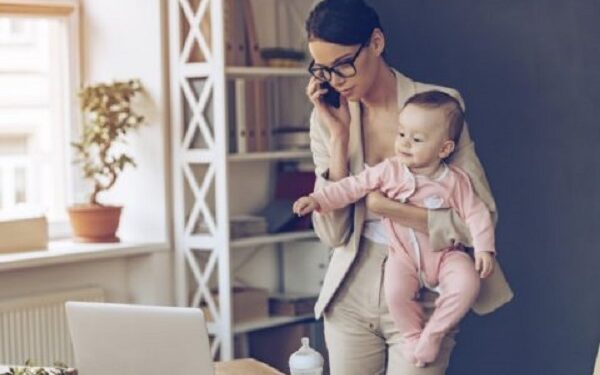 Young beautiful businesswoman talking on mobile phone and looking at laptop while standing with her baby girl at her working place