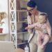 Young beautiful businesswoman talking on mobile phone and looking at laptop while standing with her baby girl at her working place