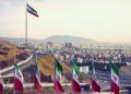 Set of Iran flags in Front of Tehran Skyline and one large flag in the background at sunset with orange warm tone.